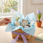 A wicker gift basket tied with a lavender ribbon and lined with light blue tissue paper, filled with bath or body products including a botanical soap packet and a lavender jar. A hand is placing white flowers into a small white vase inside the basket.