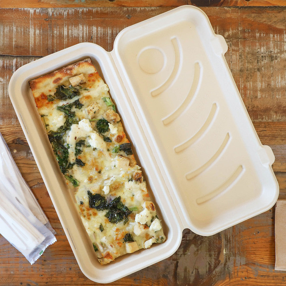 A rectangular food box with a hinged lid, shown open and filled with a baked pasta dish on a rustic wooden table. A small folded napkin or paper item is visible at the lower left.
