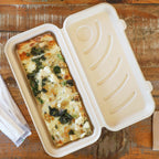 A rectangular food box with a hinged lid, shown open and filled with a baked pasta dish on a rustic wooden table. A small folded napkin or paper item is visible at the lower left.