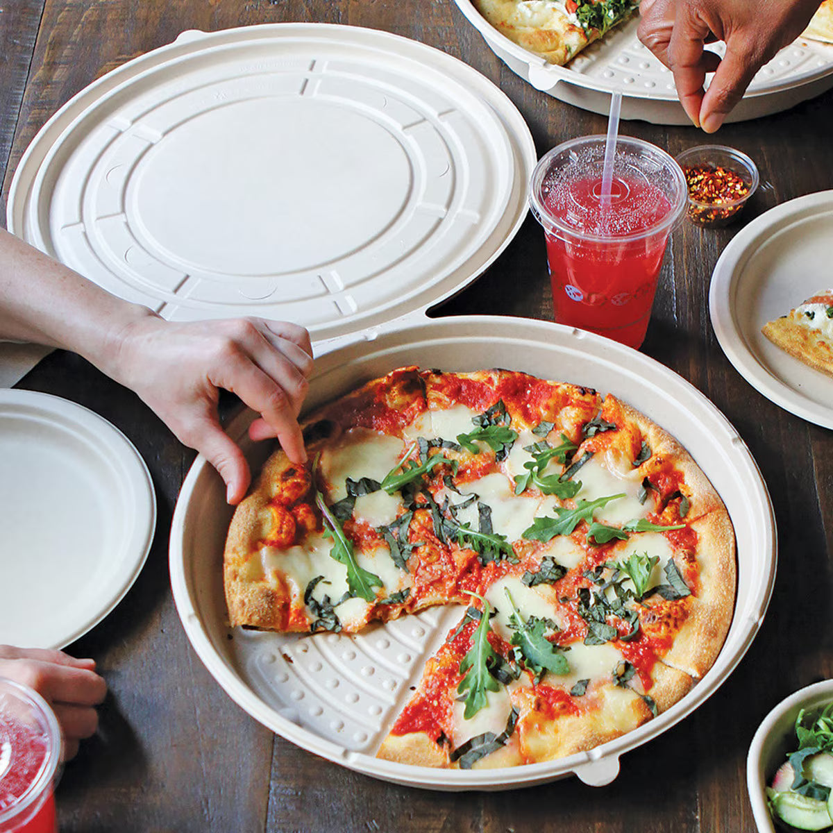 Person taking a slice of pizza from a takeout pizza boxon a wooden table with drinks and plates.