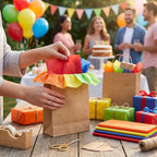 A person's hands are shown with a brown paper gift bag with bright rainbow-colored tissue paper at a festive birthday party. Other colorful gift boxes, bags, and wrapping supplies are arranged on a wooden table in the foreground, with blurred people and balloons in the background.