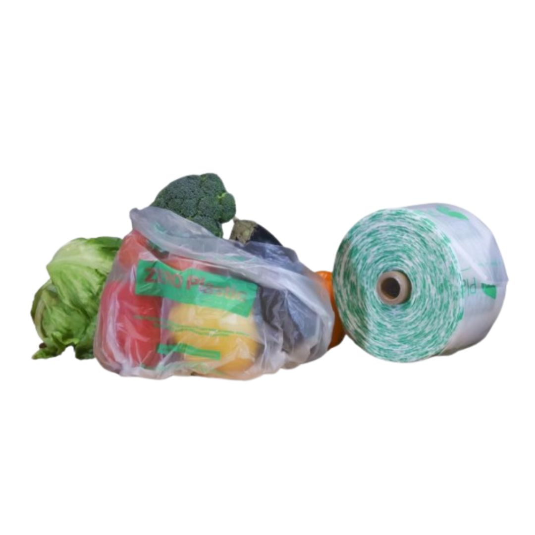 A roll of compostable plastic produce bags with green printed text, shown next to several vegetables (lettuce, broccoli, red and yellow produce) that are partially wrapped in the bags, on a plain white background.