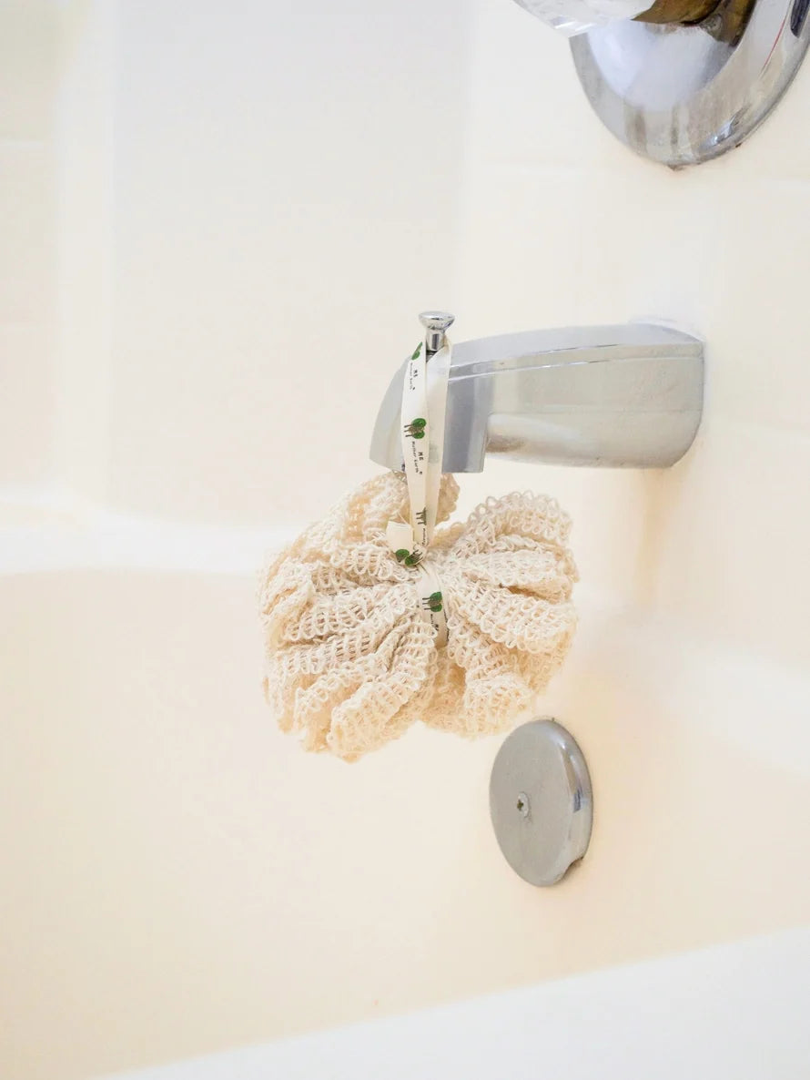 A cream-colored crochet bath sponge hanging from a chrome faucet on a white bathtub wall.