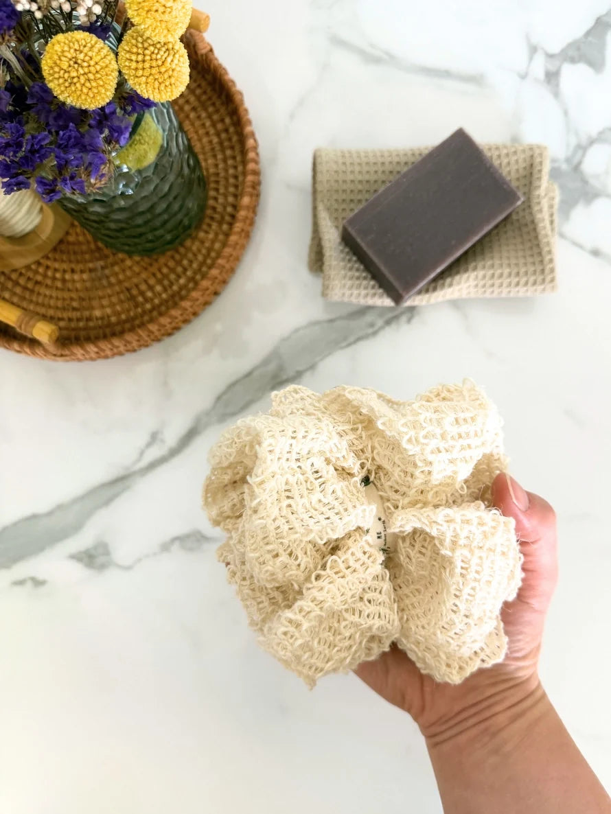 Hand holding a loofah sponge with soap bar and flowers on marble surface