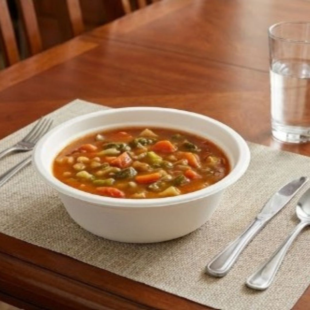 Compostable bowl of vegetable soup on a wooden table with a glass of water and cutlery.