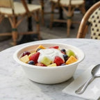 Fruit salad with yogurt in a white bowl on a marble table with chairs in the background