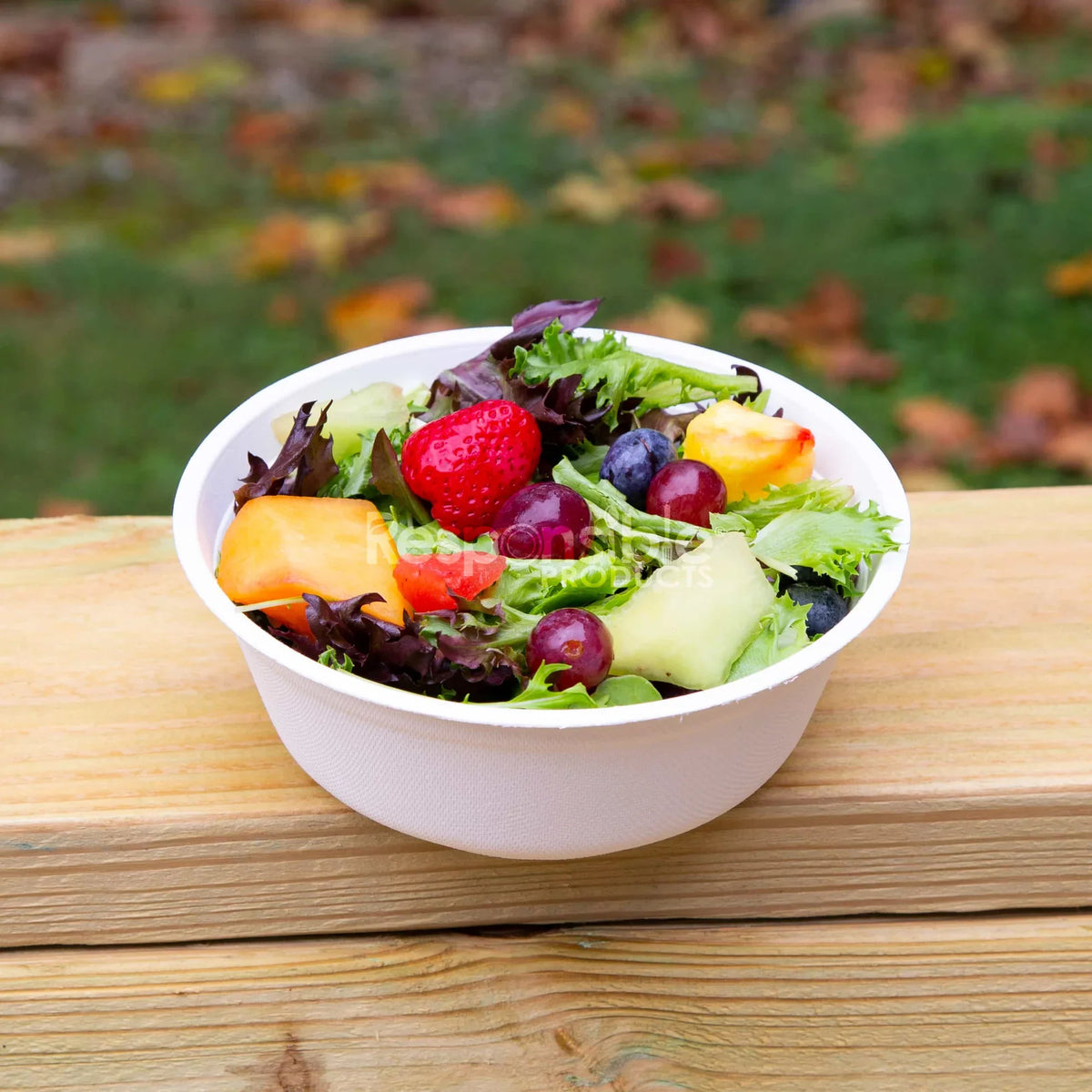 Picture of a white compostable bowl containing a salad with fruit on top of a wooden surface.