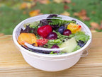 Salad with fruits and vegetables in a white bowl with a plastic lid on a wooden surface.