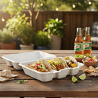 Tacos in a white taco tray with lid on a wooden table with beer bottles and condiments in the background.