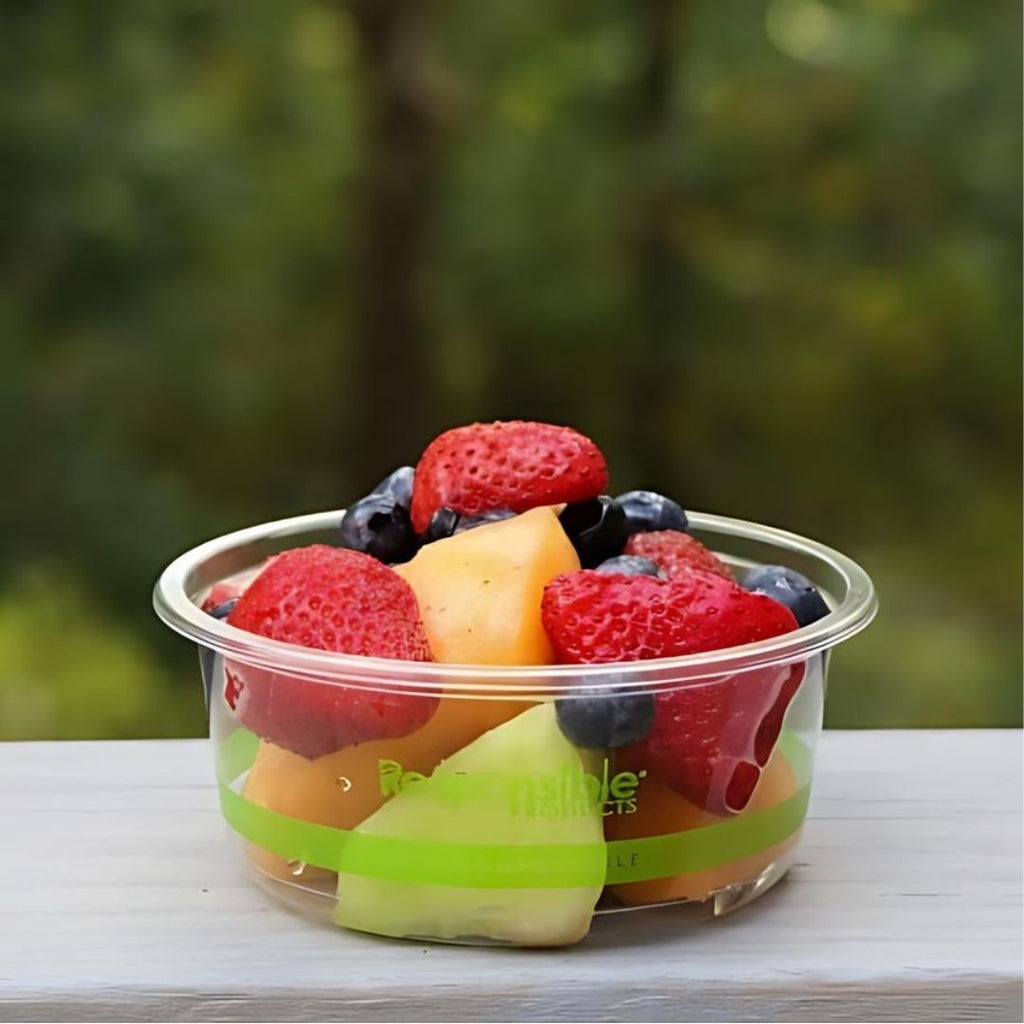 Clear deli container of mixed fruit, including strawberries, blueberries, and cantaloupe, on a blurred green outdoor background.