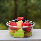 Clear deli container of mixed fruit, including strawberries, blueberries, and cantaloupe, on a blurred green outdoor background.