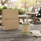 Brown paper bag and cup with branding on a wooden table outdoors.
