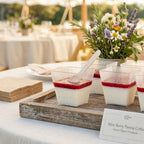 Tray of mini berry panna cotta in compostable dessert containers with a floral arrangement on a table outdoors.