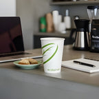 Compostable cup with green logo on a desk with laptop, cookies, and notebook.