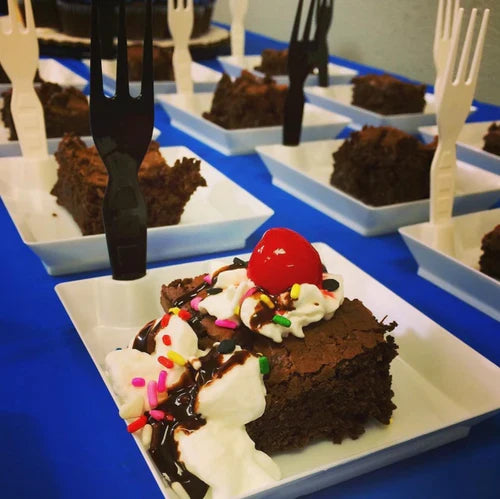 Chocolate brownie dessert with whipped cream and a cherry on top, served with on a square white plate.