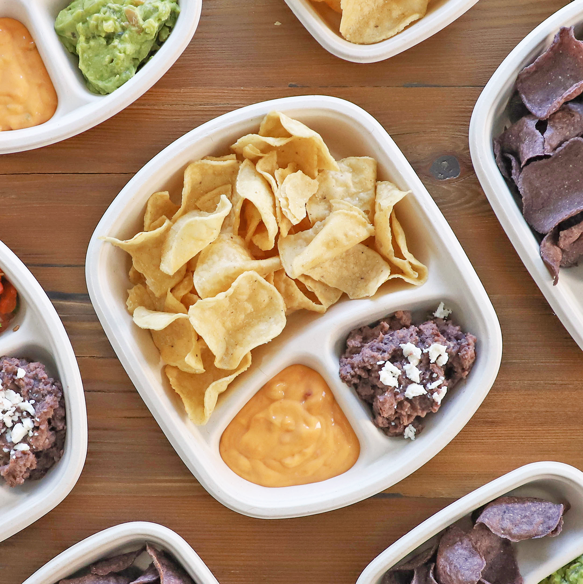 Three Compartment Tray containing chips, nacho cheese, and beans next to more trays on a wooden table.