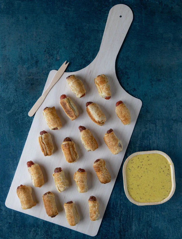 Baked sausage rolls on a white cutting board with a side of green sauce on a blue background