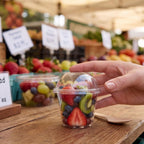 A hand holds a compostable portion cup with lid filled with mixed fruit, including strawberries, blueberries, and kiwi, on a wooden table at an outdoor market.