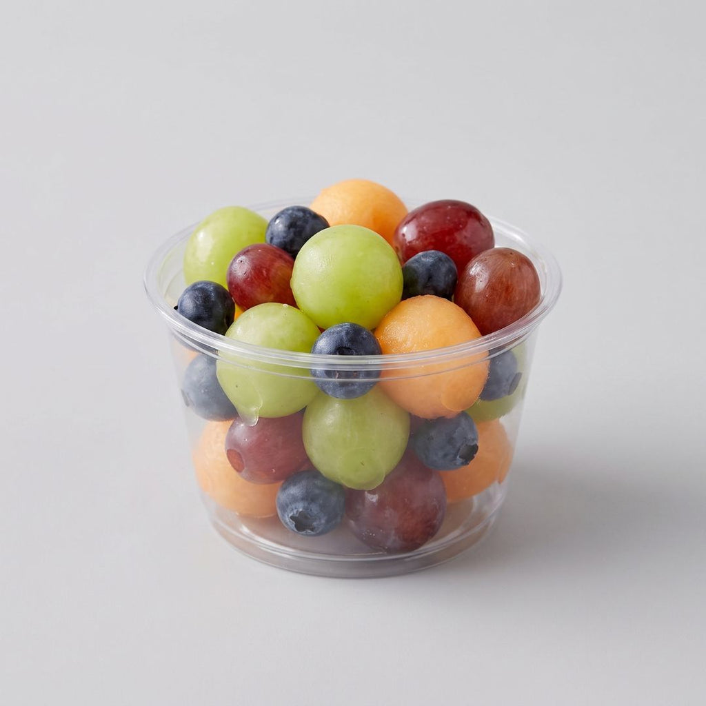 A clear compostable side cup filled with assorted fresh fruit, including green grapes, blueberries, red grapes, and melon balls, on a light gray background.