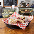 A cut-open sandwich with sliced meats and cheese sits on red-and-white checkered paper inside a dark woven basket, alongside a small clear cup of coleslaw, on a wooden table in a restaurant setting.