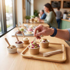 A close-up of a hand holding a wooden spoon and eating a scoop of colorful berry ice cream from a small paper cup on a wooden tray, with other ice cream cups and blurred people in a cafe background.