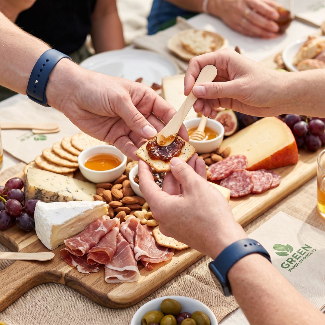 A close-up lifestyle shot of a charcuterie board being shared by multiple people. Hands are shown spreading jam on a piece of bread using a compostable bowtie spoon, with cheeses, crackers, nuts, salami, grapes, and small bowls of spreads and olives arranged on a wooden board.