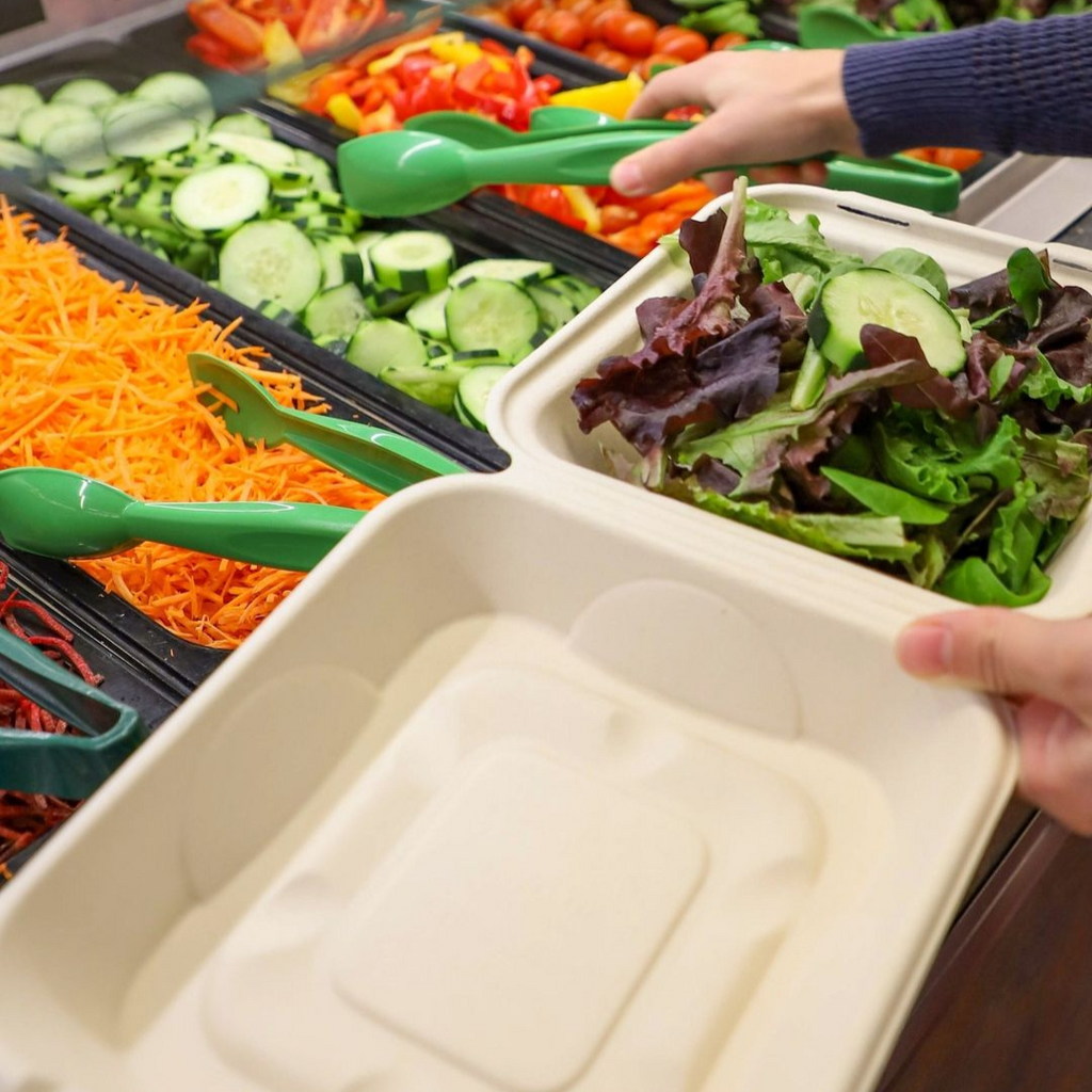 Person at the salad bar, using a green utensil to fill a compostable clamshell with salad.