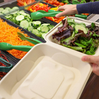 Person at the salad bar, using a green utensil to fill a compostable clamshell with salad.