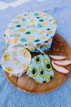 Fruit-themed wax-wrapped foods on a wooden plate with apple slices on a blue patterned background