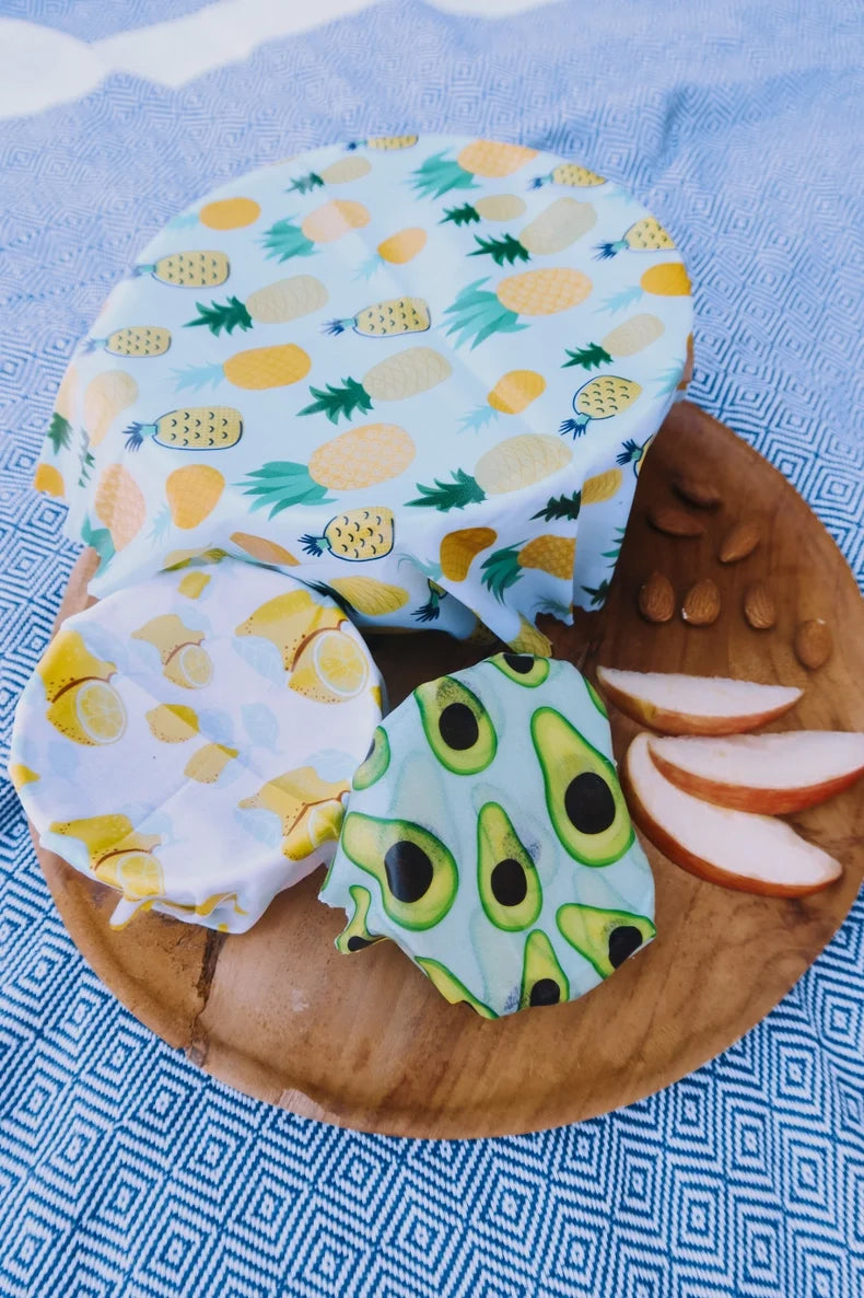 Fruit-themed wax-wrapped foods on a wooden plate with apple slices on a blue patterned background