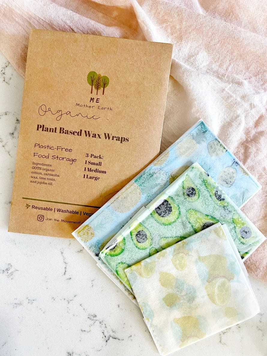A package of organic plant-based wax wraps lies on a marble surface next to three reusable cloth wraps with avocado and lime patterns, all styled with a soft pink fabric backdrop.
