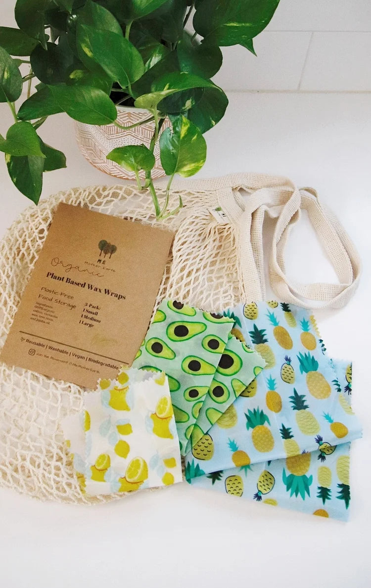Set of reusable food wraps with avocado and pineapple patterns on a white surface with a plant in the background.