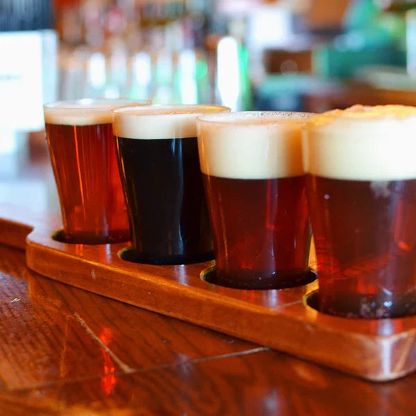 Four compostable glasses of different types of beer on a wooden tray with a blurred bar background.