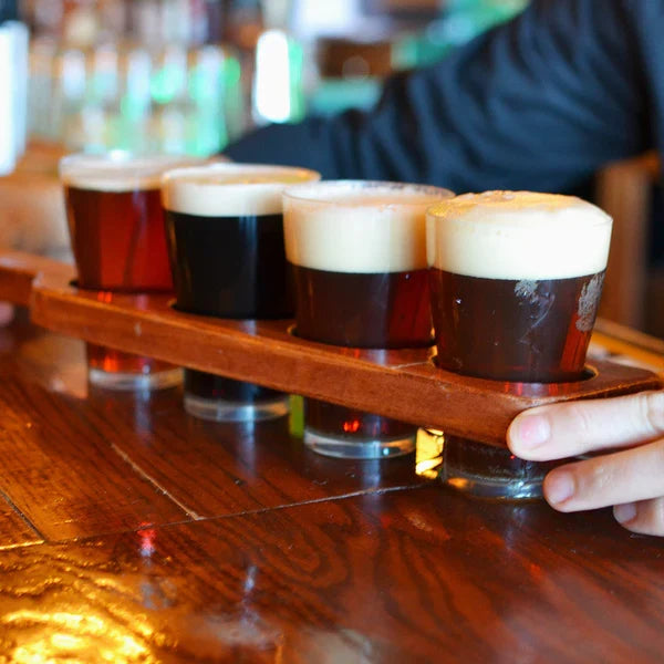 Four beer samples served in compostable beer cups on a wooden tray held by a person in a bar setting.