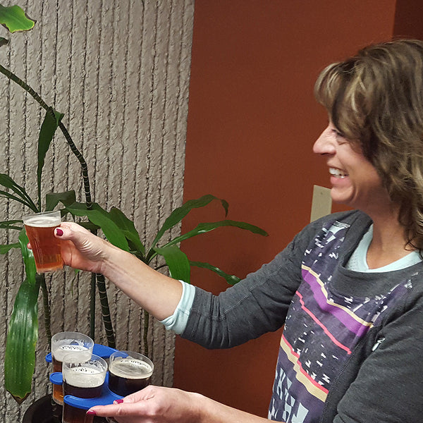 Woman holding a compostable cup of beer next to a tray with more glasses indoors.