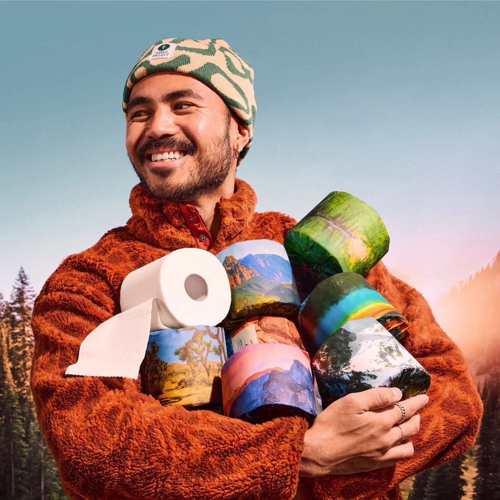 A smiling man wearing a fuzzy orange-brown fleece jacket and a green camouflage beanie holds several toilet paper rolls printed with colorful national park landscape scenes against a forest and sky background.