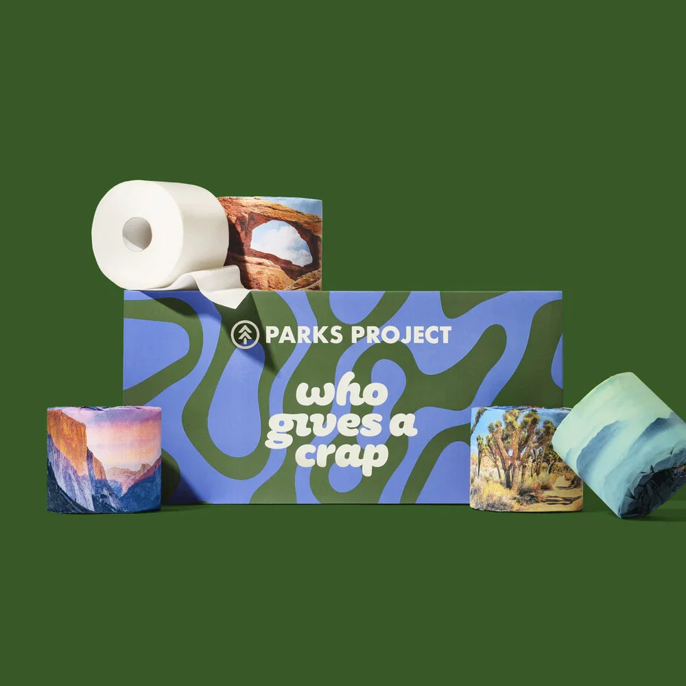 Parks Project toilet paper and tissue products arranged on a dark green background, including a roll of white toilet paper, a blue box with the brand name and slogan, and several tissue packs with landscape prints.