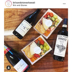 Three bottles of wine with trays of food on a wooden table, featuring the brand 'Brix and Stones'.