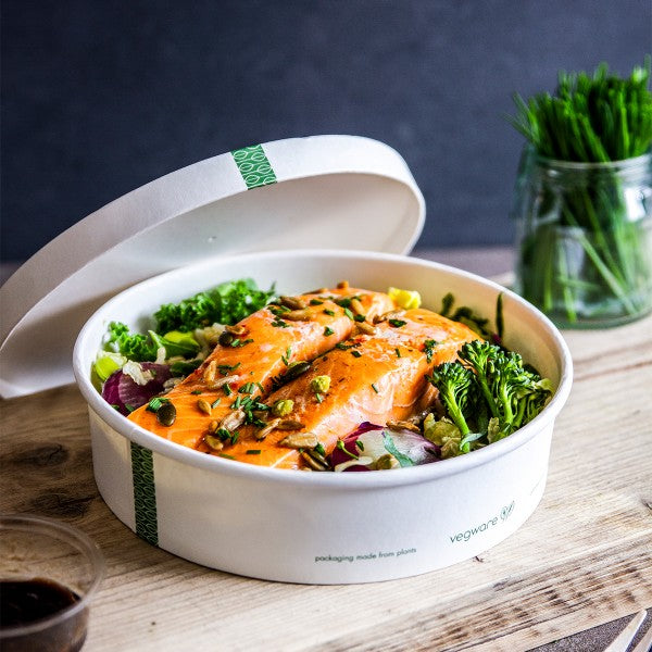 Compostable white bowl containing salmon and salad with a compostable bowl lid resting on the bowl on a wooden table.