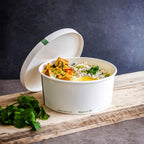 Compostable white paper bowl filled with food with lid resting near by sitting on a wooden table with a blue background.