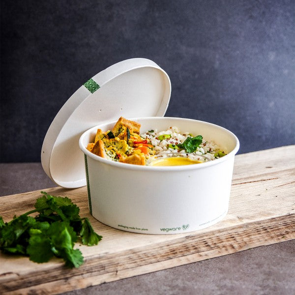 Compostable white paper bowl filled with food with lid resting near by sitting on a wooden table with a blue background.