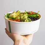 Compostable paper bowl containing salad that is being held by a hand with a plain white background.