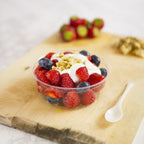 A compostable round deli container filled with berries and yogurt with a spoon resting on a cutting board with a blurred background.