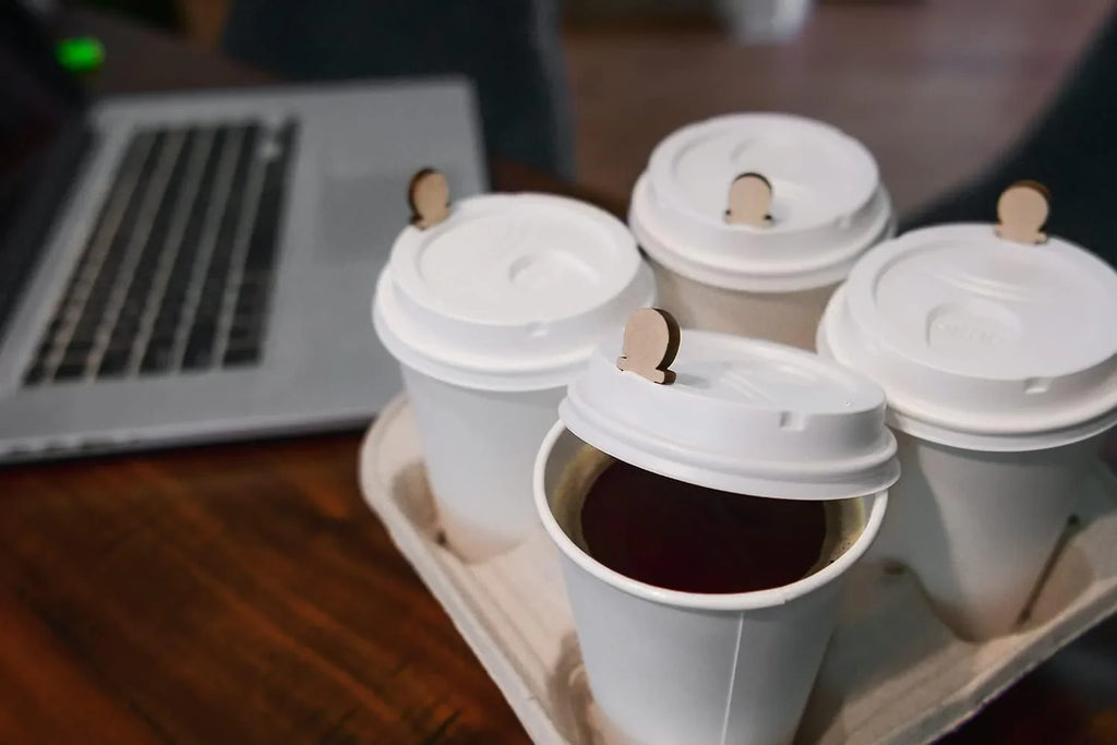 Four paper coffee cups with lids containing wooden lid plugs, in a cup carrier, next to a laptop on a wooden table.