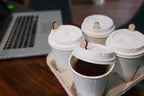 Four paper coffee cups with lids containing wooden lid plugs, in a cup carrier, next to a laptop on a wooden table.